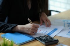 Female executives are finalizing a budget plan. She uses the pen to point to the document. Calculator and note book on the desk.